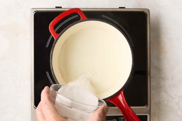Sugar being added to cream in a saucepan over a burner.