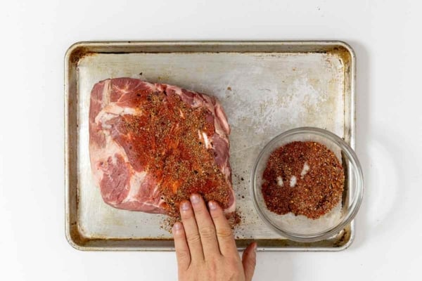 A man's hand rubbing spice mix onto a pork butt or pork shoulder.