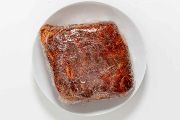A pork butt wrapped in plastic on a white plate.