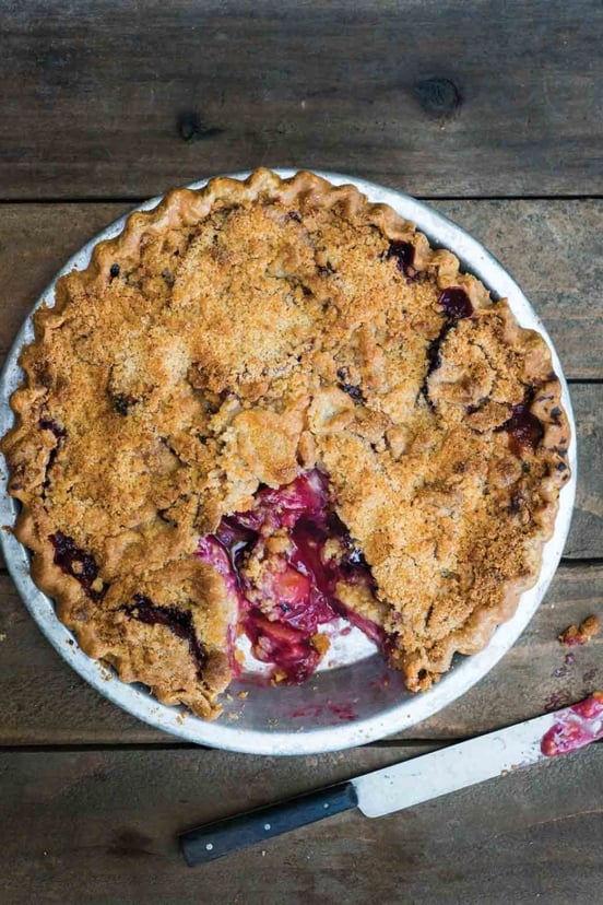 A plum pie topped with crumble with one piece missing from the pie.