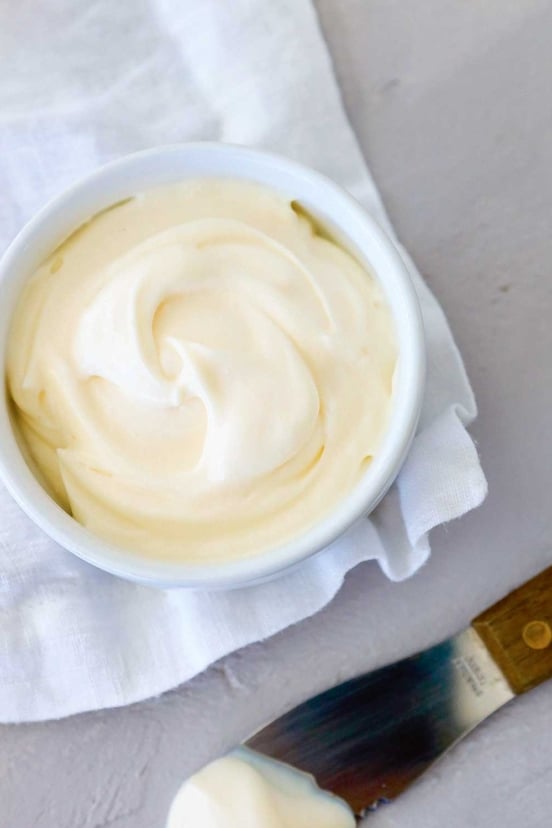 A bowl of homemade mayonnaise on a white cloth with a knife on the side.