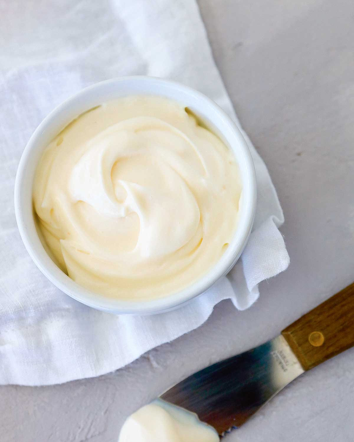 A bowl of homemade mayonnaise on a white cloth with a knife on the side.