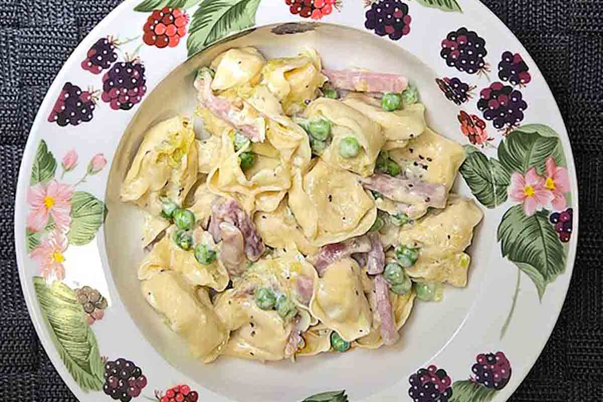 A plate filled with tortellini in a creamy sauce with peas and prosciutto.