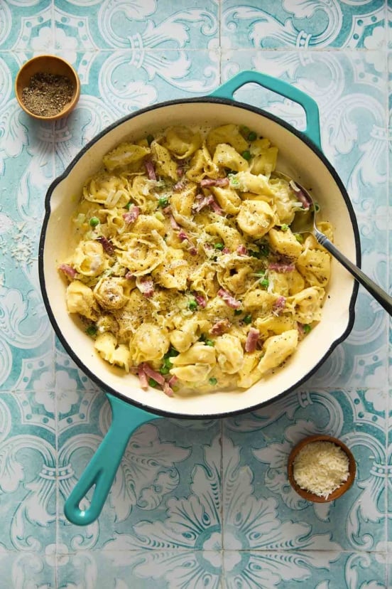 A deep skillet filled with tortellini with peas and prosciutto with bowls of pepper and cheese on the side.