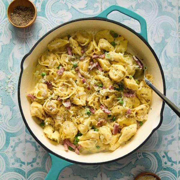 A deep skillet filled with tortellini with peas and prosciutto with bowls of pepper and cheese on the side.