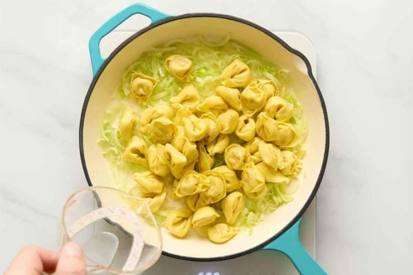 Tortellini and water being added to a skillet with leeks.