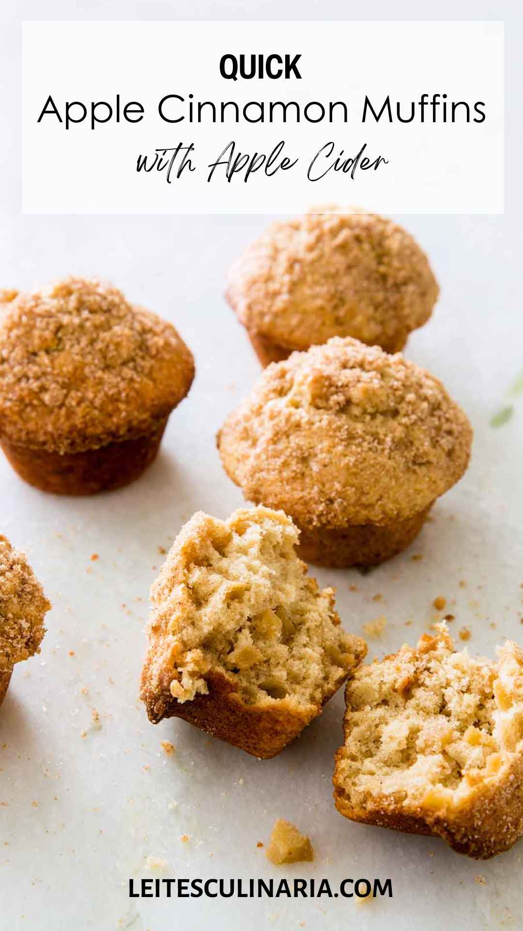 Sugar-topped apple cinnamon muffins on a white counter. The muffin in the forefront is broken in half.