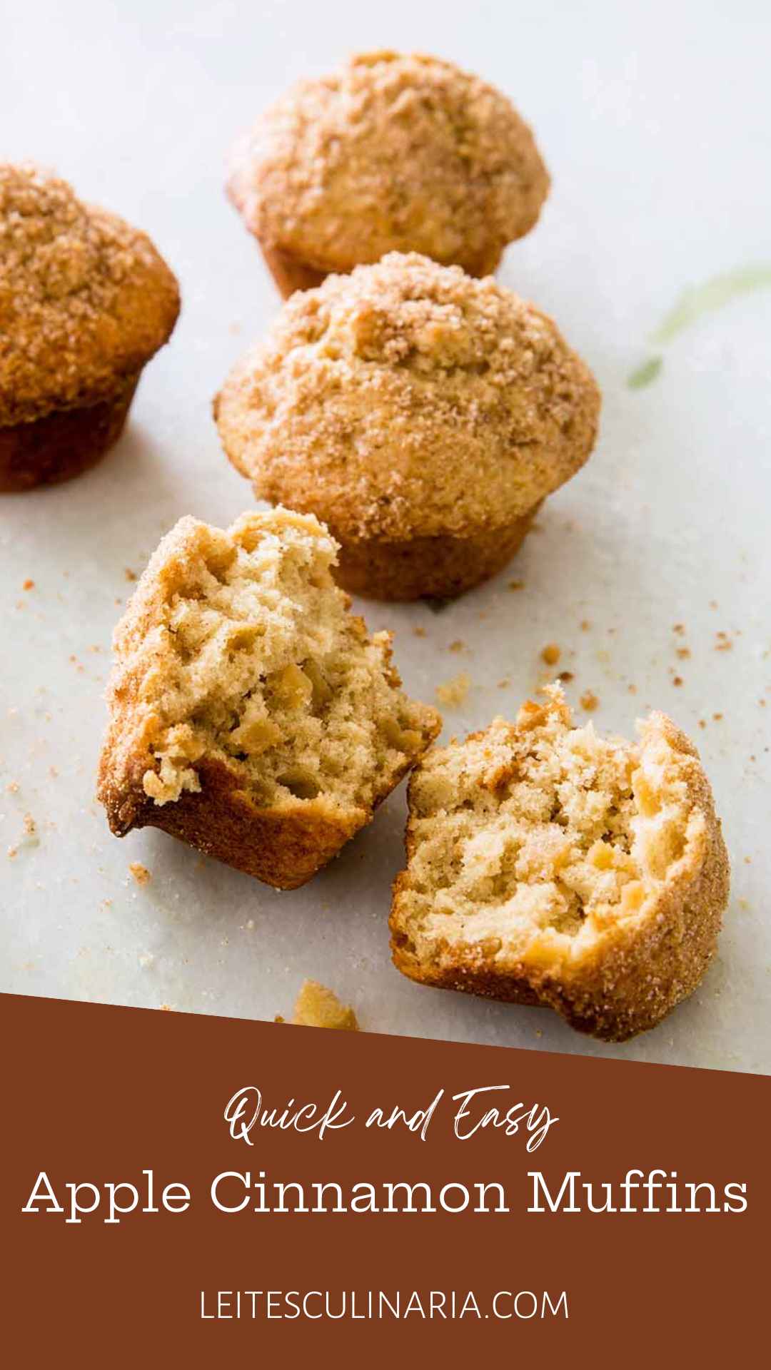 Sugar-topped apple cinnamon muffins on a white counter. The muffin in the forefront is broken in half.