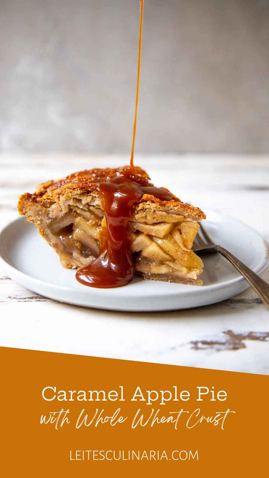 A slice of homemade caramel apple pie on a white plate. A thin stream of homemade caramel is drizzling over the top crust of the pie. A fork sits on the right next to the pie.