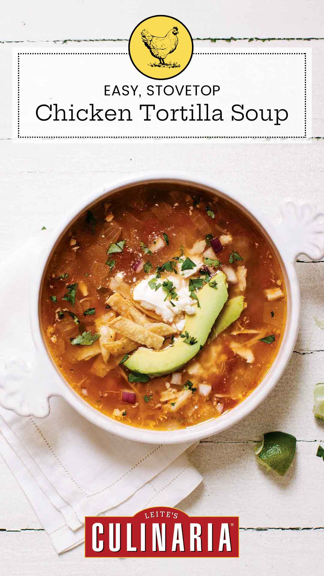 A white crock of chicken tortilla soup topped with avocado slices, tortilla strips and a dollop of sour cream. A quartered lime is setting in the lower right near the crock.