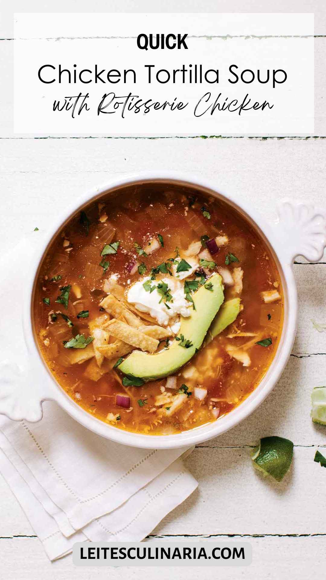 A white crock of chicken tortilla soup topped with avocado slices, tortilla strips and a dollop of sour cream. A quartered lime is setting in the lower right near the crock.