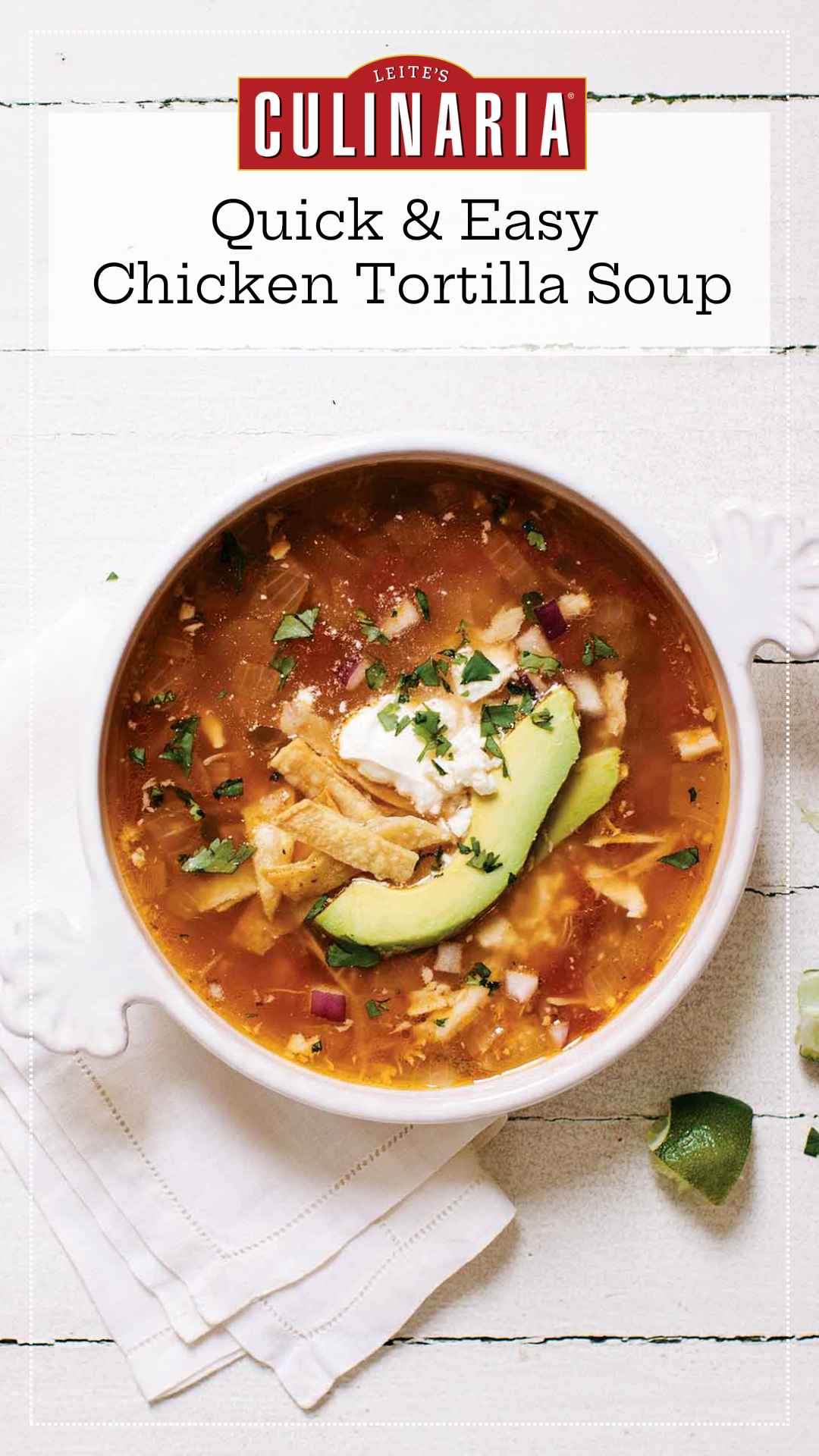 A white crock of chicken tortilla soup topped with avocado slices, tortilla strips and a dollop of sour cream. A quartered lime is setting in the lower right near the crock.