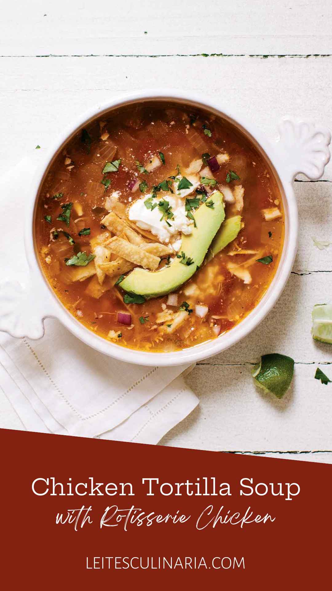 A white crock of chicken tortilla soup topped with avocado slices, tortilla strips and a dollop of sour cream. A quartered lime is setting in the lower right near the crock.