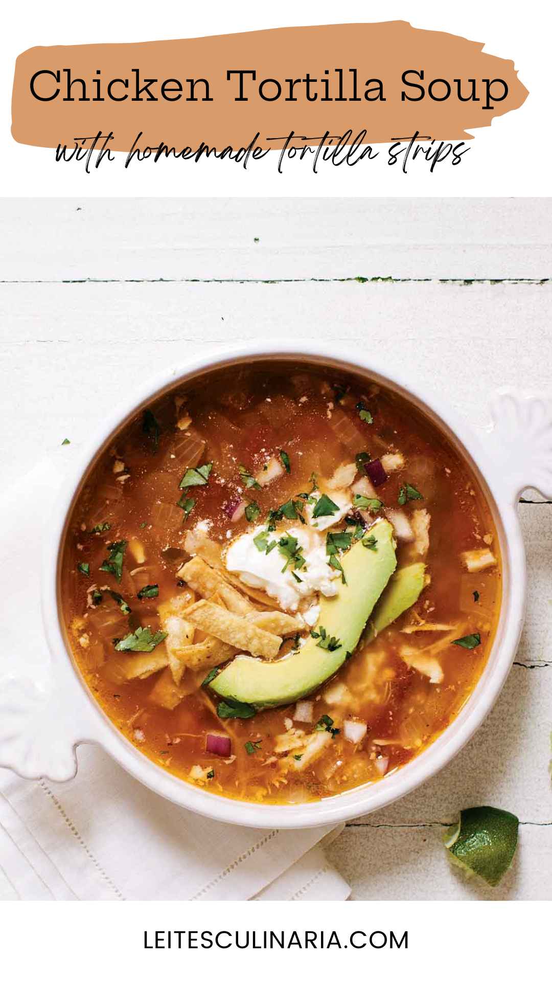 A white crock of chicken tortilla soup topped with avocado slices, tortilla strips and a dollop of sour cream. A quartered lime is setting in the lower right near the crock.