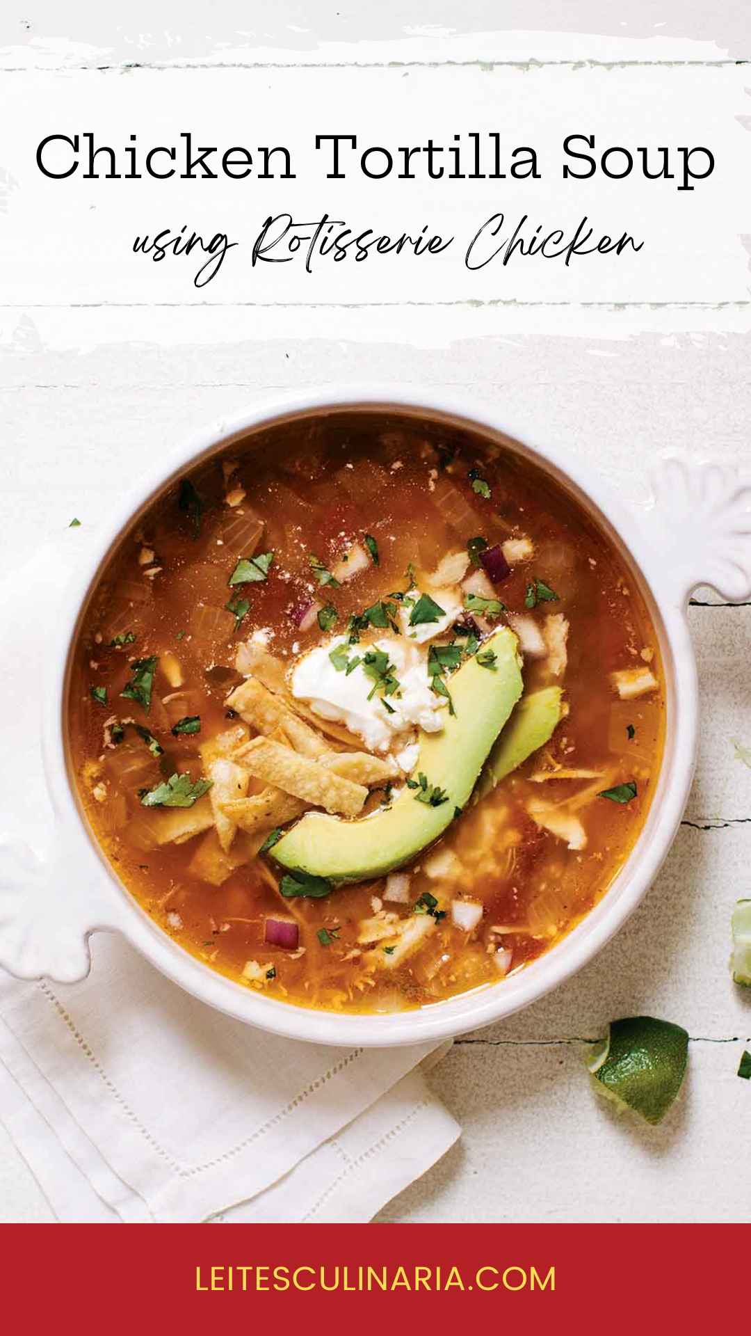 A white crock of chicken tortilla soup topped with avocado slices, tortilla strips and a dollop of sour cream. A quartered lime is setting in the lower right near the crock.