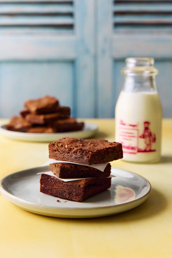 Three brownies stacked on a plate with a bottle of milk in the background.