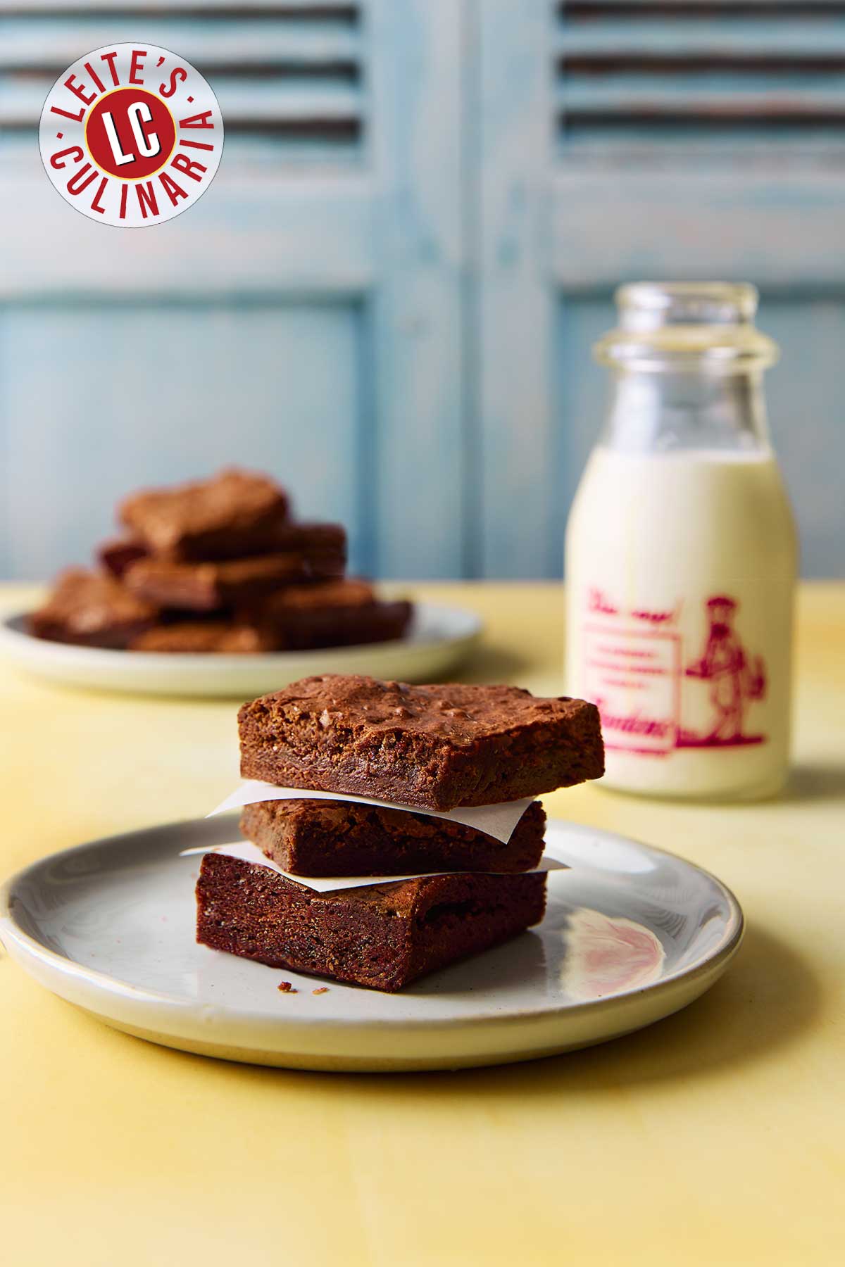 Three brownies stacked on a plate with a bottle of milk in the background.