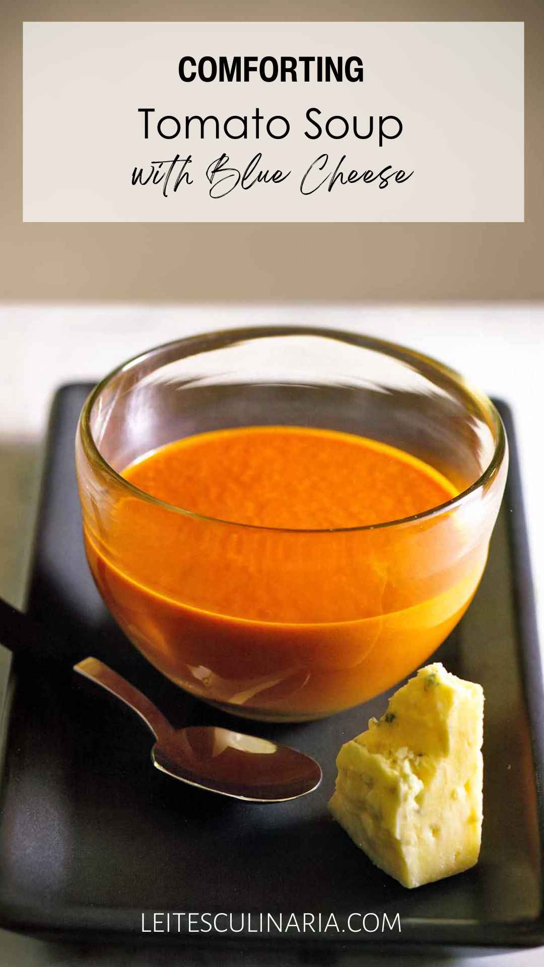 A bowl of tomato soup with a spoon and a wedge of blue cheese on the side.