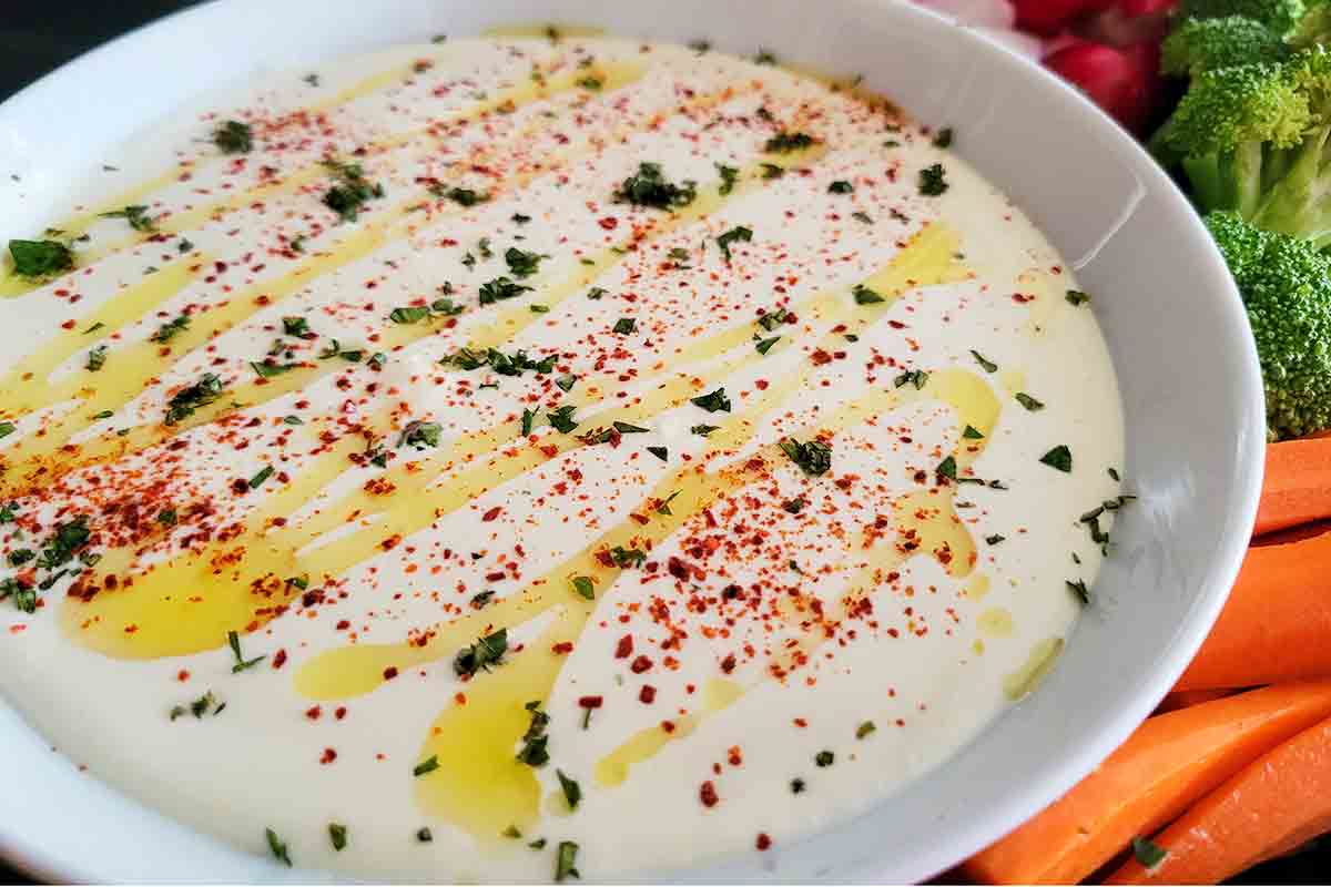 A white bowl filled with feta dip drizzled with oil and topped with spices, with raw veggies on the side.