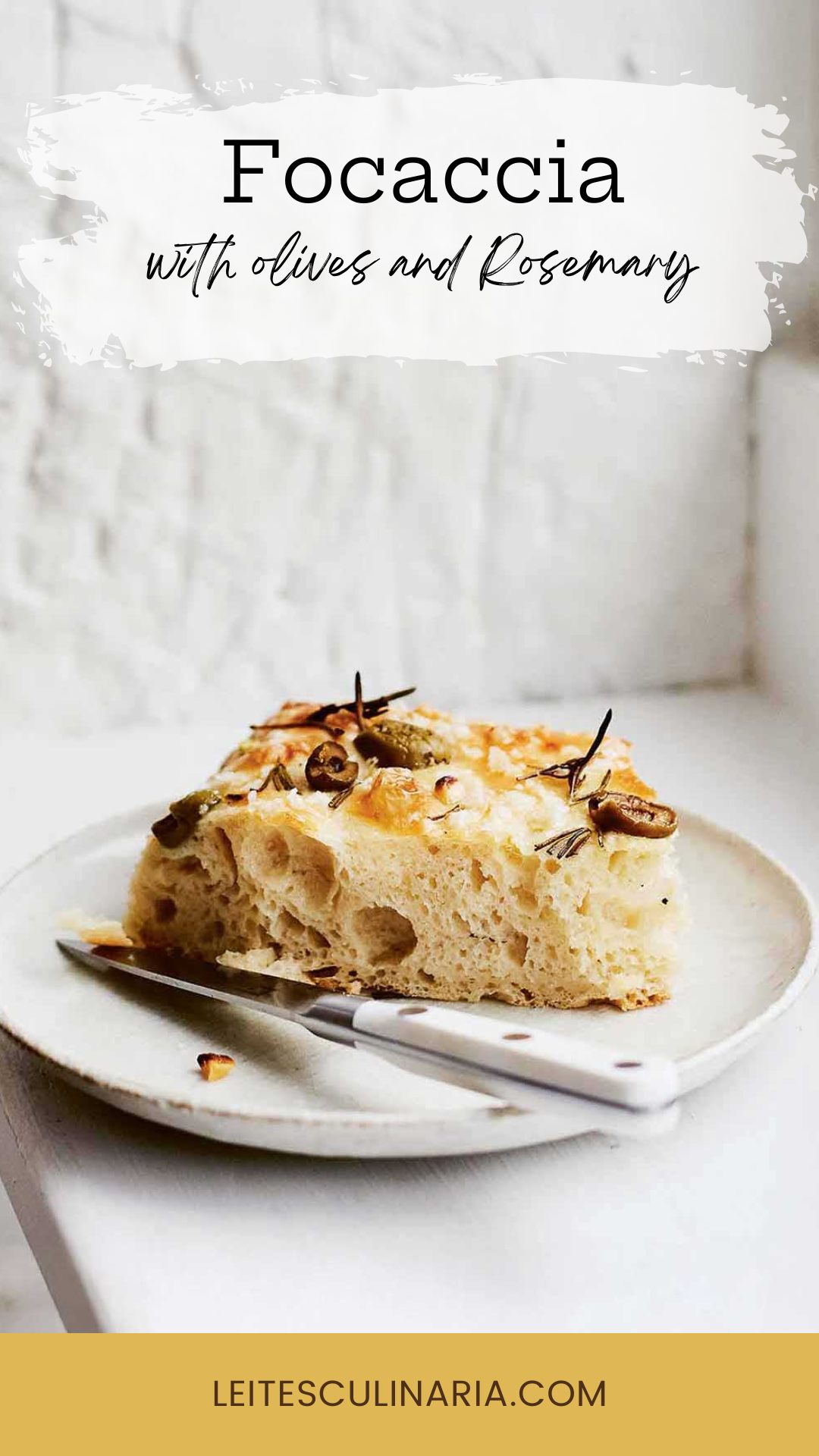 A slice of focaccia with olives and rosemary on a white plate with a knife resting beside it.