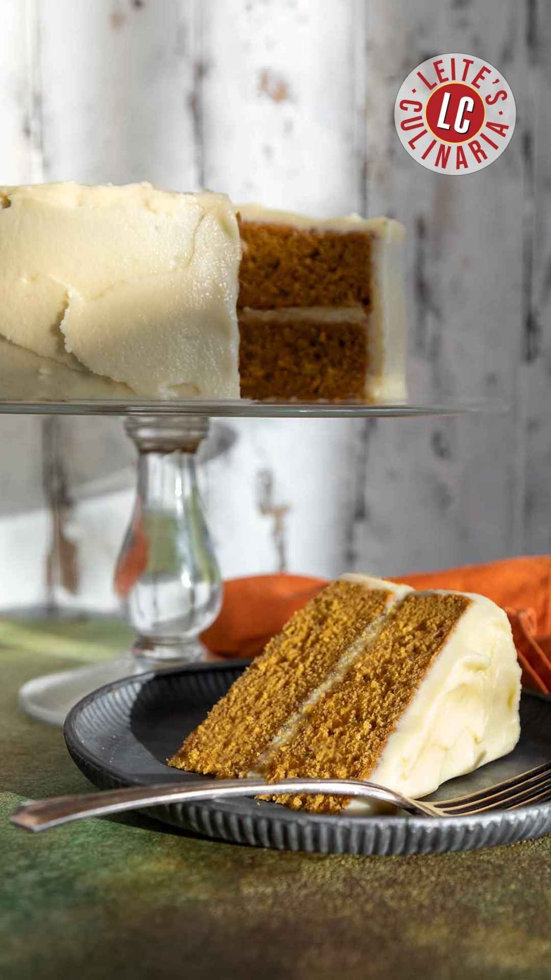 A slice of frosted pumpkin cake on a plate with the remaining cake on a stand in the background.