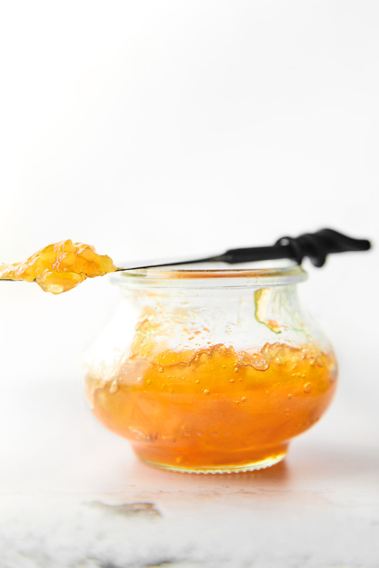 A small jam jar filled with Meyer lemon marmalade. Resting on top is a knife with a dollop of marmalade on top.