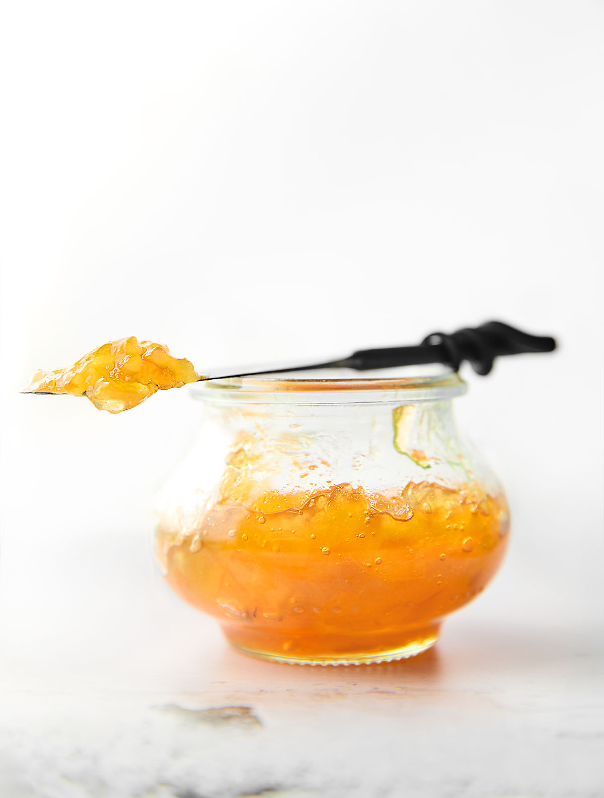 A small jam jar filled with Meyer lemon marmalade. Resting on top is a knife with a dollop of marmalade on top.