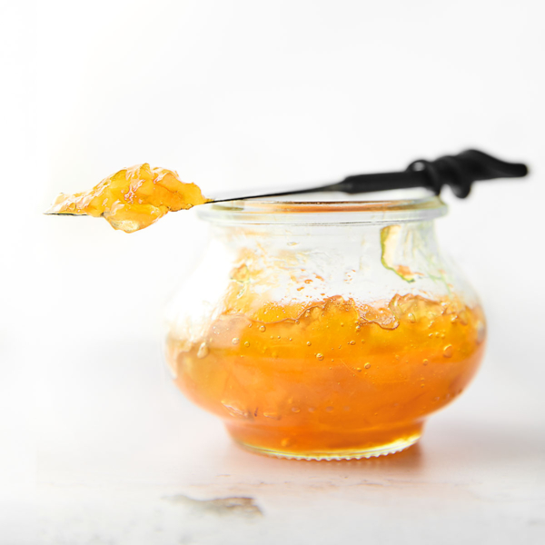 A small jam jar filled with Meyer lemon marmalade. Resting on top is a knife with a dollop of marmalade on top.