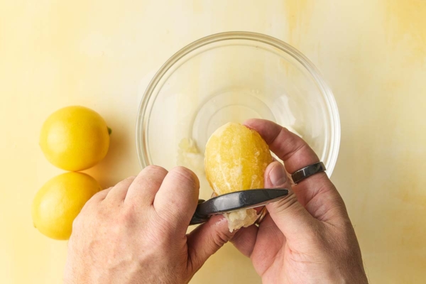 A man's hand peeling a Meyer lemon with a vegetable peeler over a glass bowl.