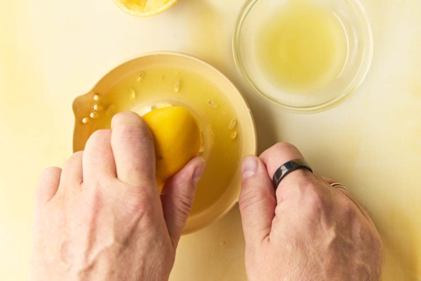 A man's handing juicing a Meyer lemon half. Nearby is a glass bowl of lemon juice.