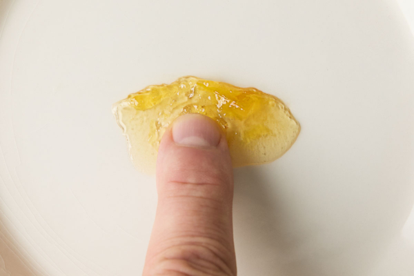 A fingertip gently pressing a small dollop of Meyer lemon marmalade on a white plate to test its consistency.