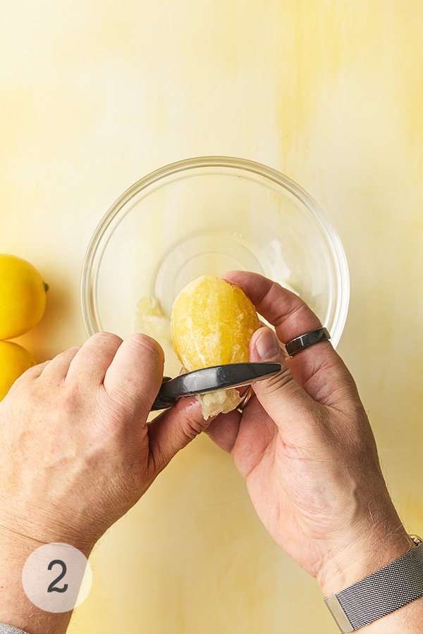 A man's hand peeling a Meyer lemon with a vegetable peeler over a glass bowl. 