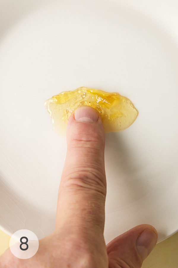 A fingertip gently pressing a small dollop of Meyer lemon marmalade on a white plate to test its consistency.