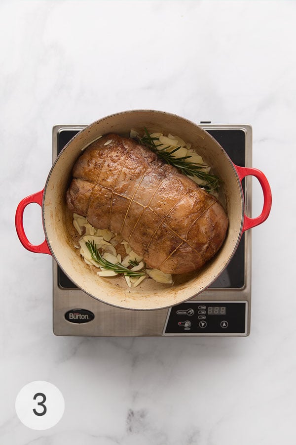 A seared leg of lamb in a Dutch oven with garlic slivers and fresh rosemary.