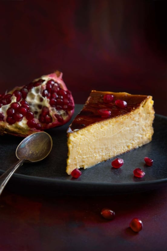 A slice of burnt Basque cheesecake with pomegranate on a plate, garnished with pomegranate seeds and a spoon.