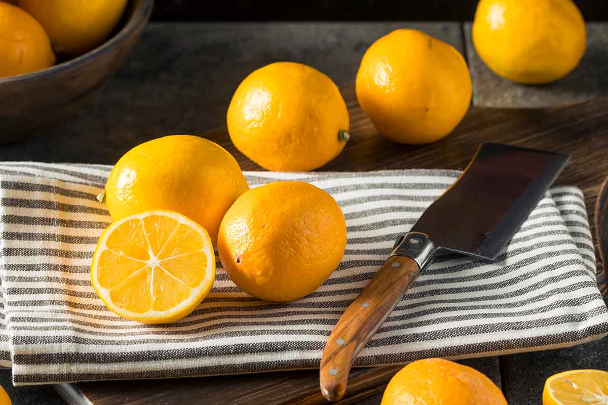 Several Meyer lemona on a striped towel; one is sliced in half. Nearby is a cleaver.