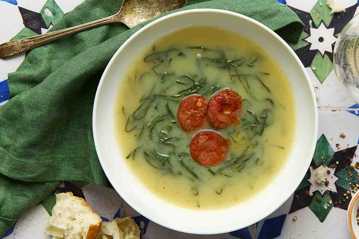 A white bowl of caldo verde, or Portuguese kale soup (green broth) with thinly sliced kale and three chourico coins floating in the center.