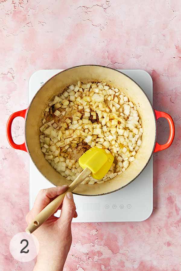 A man's hand stirring a pot of onions and garlic in a pot.