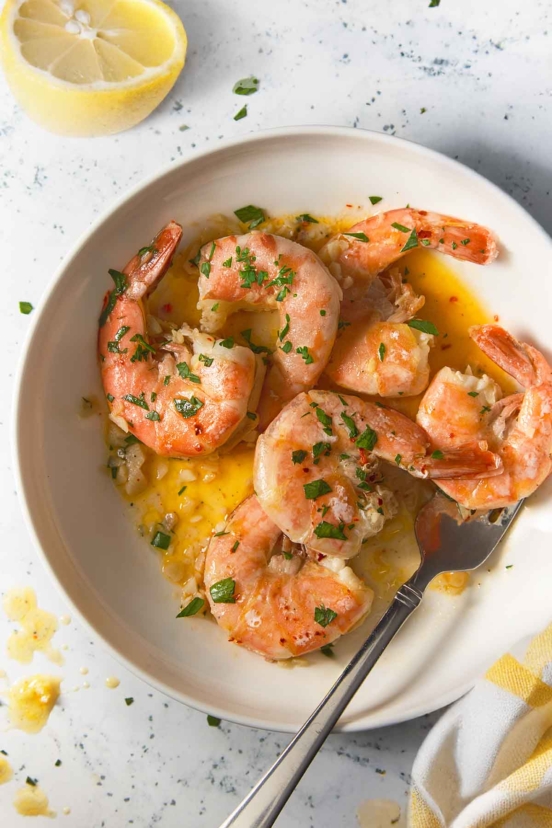 Classic shrimp scampi on a white plate with large pink shrimp in garlic butter sauce, garnished with parsley. A silver fork rests on the side, with a lemon half nearby and golden sauce pooling at the bottom for dipping.