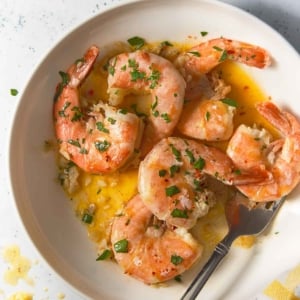 A wide shot of shrimp scampi in a white bowl, drenched in garlic butter sauce and garnished with parsley. A lemon wedge sits nearby, with scattered herbs and sauce on the table.