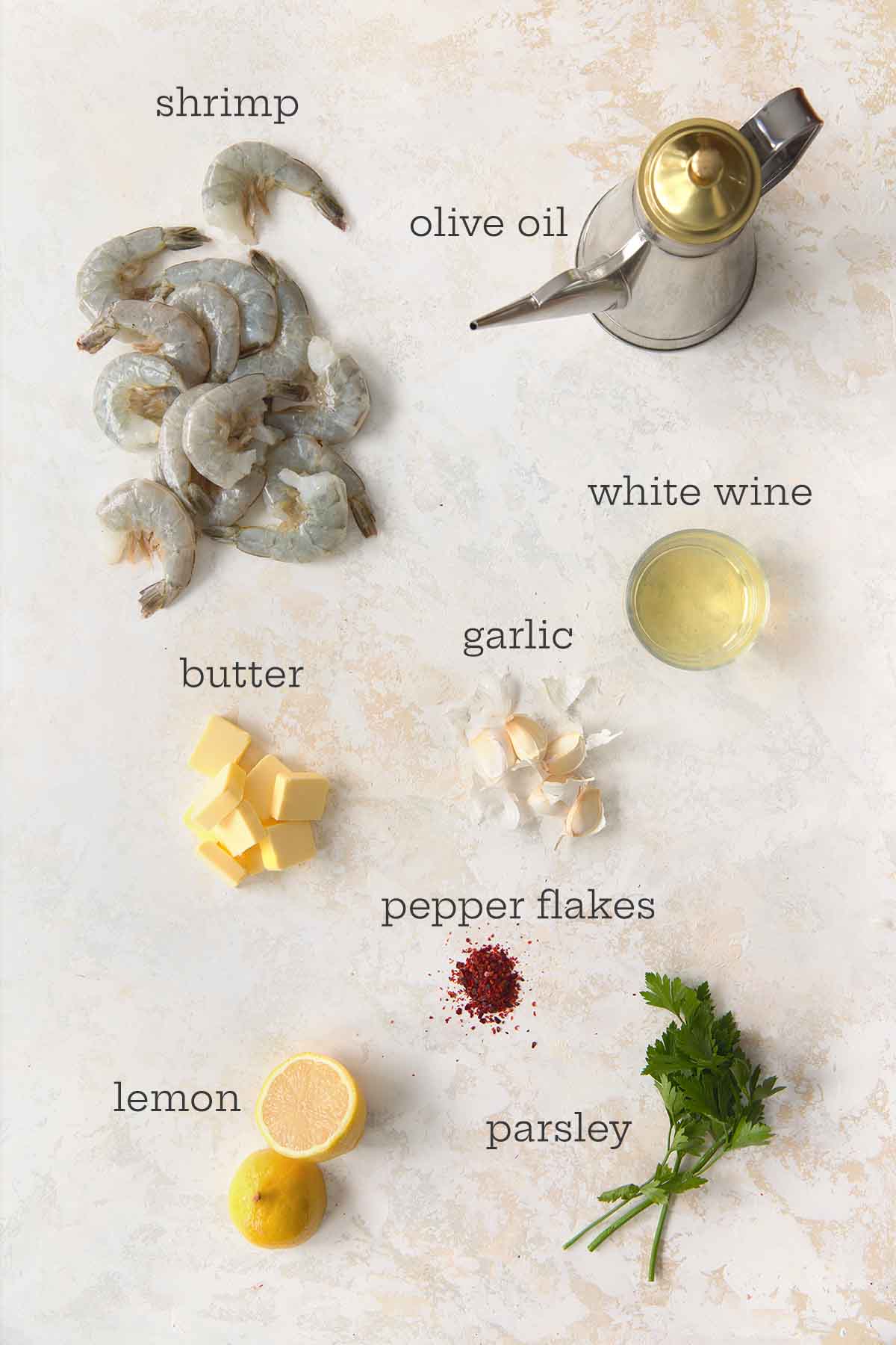 Overhead view of raw shrimp, olive oil, white wine, garlic cloves, butter cubes, red pepper flakes, lemon halves, and fresh parsley arranged on a light surface.