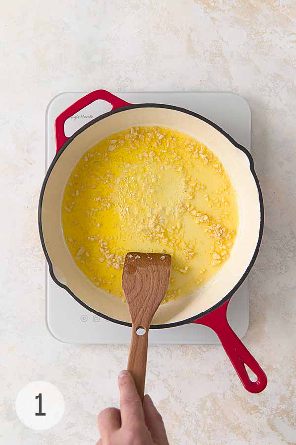 Garlic cooking in melted butter and olive oil in a red-handled skillet over medium heat, stirred with a wooden spatula.