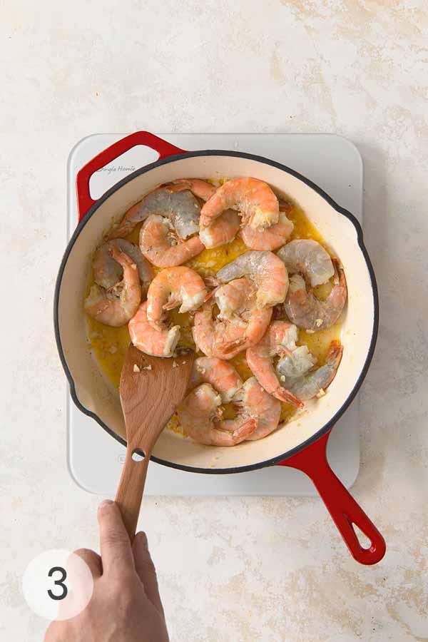 Raw shrimp cooking in a garlic butter and white wine sauce in a skillet, stirred with a wooden spatula.