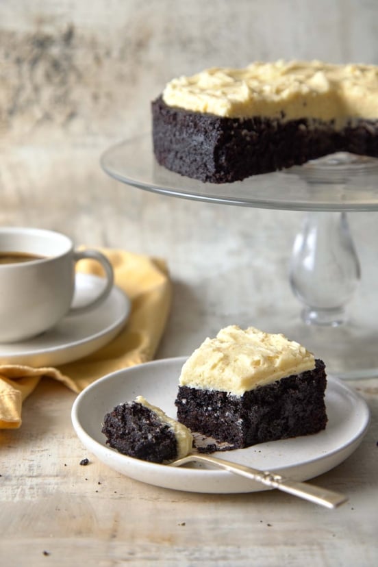 Guinness chocolate cake with browned butter frosting on a glass cake stand, with a slice on a plate and a cup of coffee in the background.