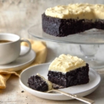 Chocolate Guinness cake with browned butter frosting on a glass cake stand, with a slice on a plate and a cup of coffee in the background.