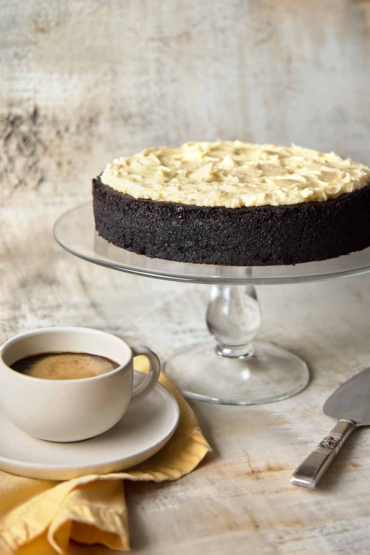 Side view of a rich, moist Guinness chocolate cake topped with browned butter cream cheese frosting displayed on a cake stand, accompanied by a cup of coffee.