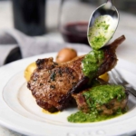 Juicy lamb chops on a white plate with a drizzle of vibrant mint and cilantro sauce.