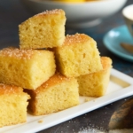 Moist lemon drizzle cake squares stacked on a white plate with a sugar crust, next to a cup of coffee and a bowl of lemons.