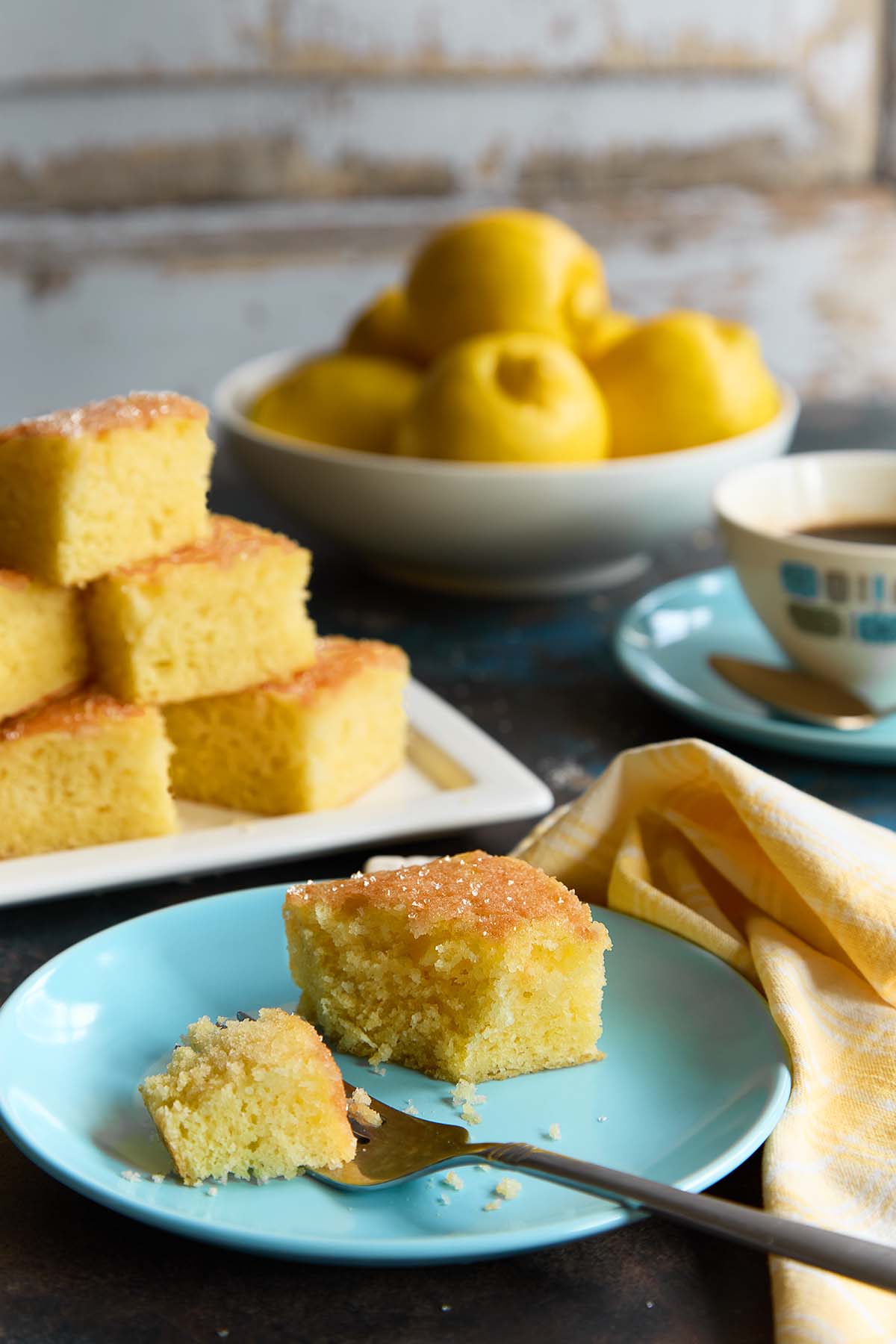 A slice of lemon drizzle cake with a sugar crust on a blue plate, fork beside it, with a bowl of lemons and a coffee cup in the background.