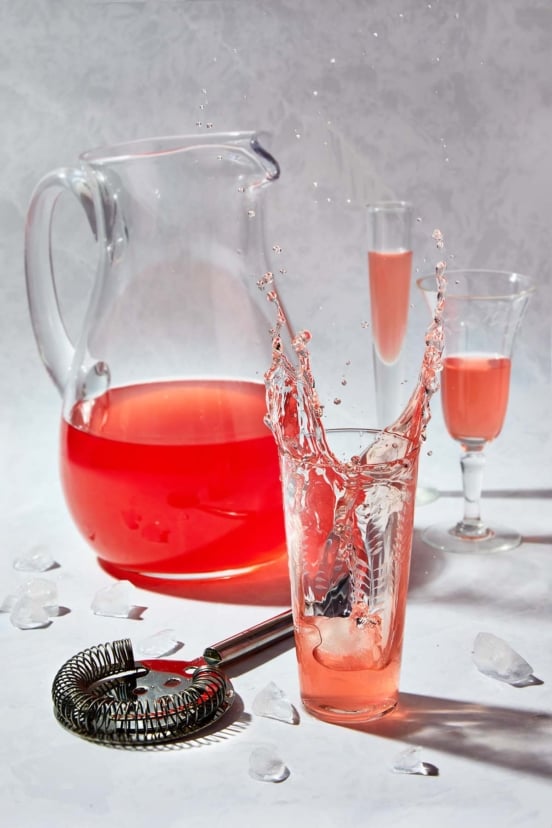 Rhubarb vodka splashing over ice beside a glass pitcher and cocktail tools.